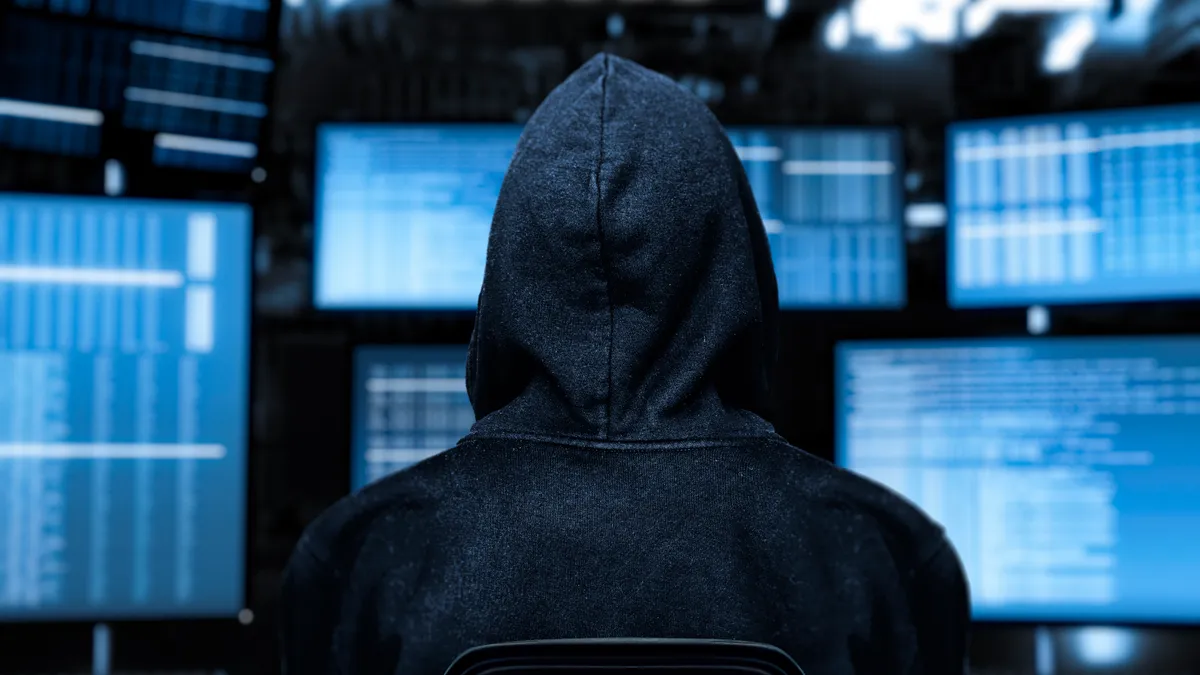 Hooded hacker sits in front of computer screens.