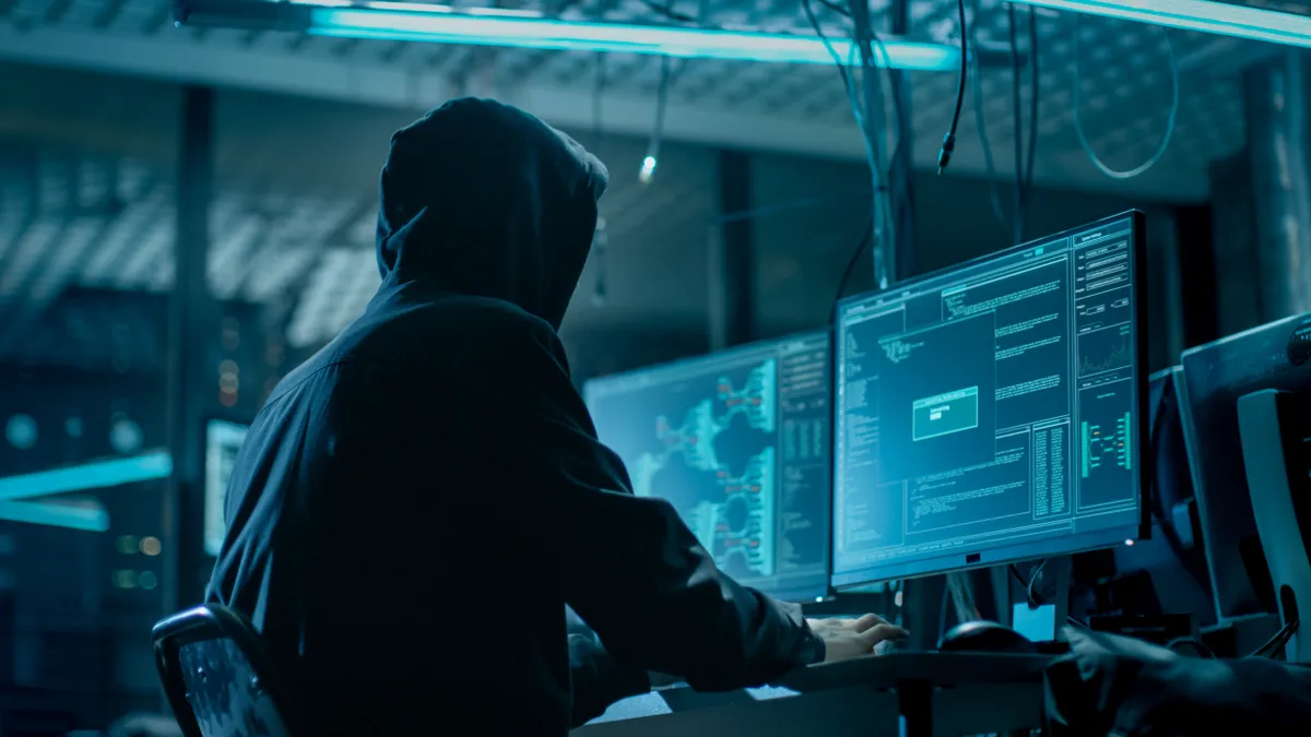 Hooded person types on computer in a dark room with multiple monitors and cables everywhere.