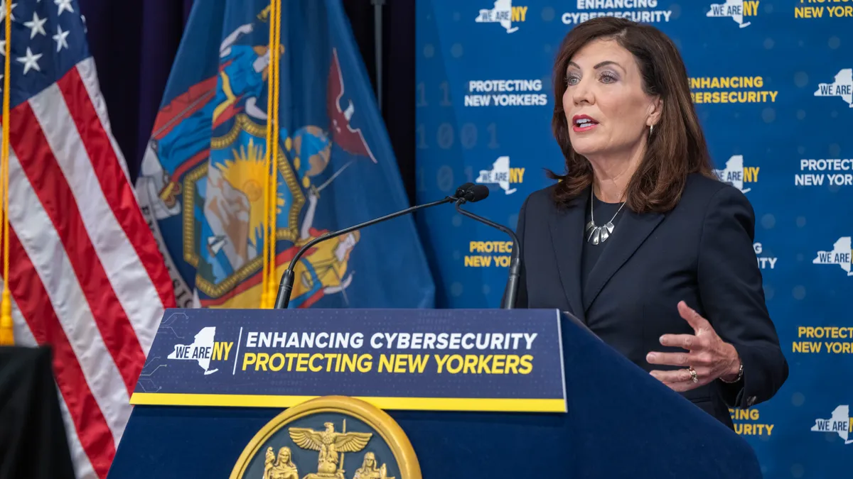Woman in a black suit stands behind a podium with a sign that reads "enhancing cybersecurity protecting New Yorkers."