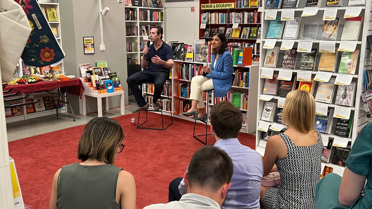 A woman speaking to a man in a bookstore in front of an audience on risers.
