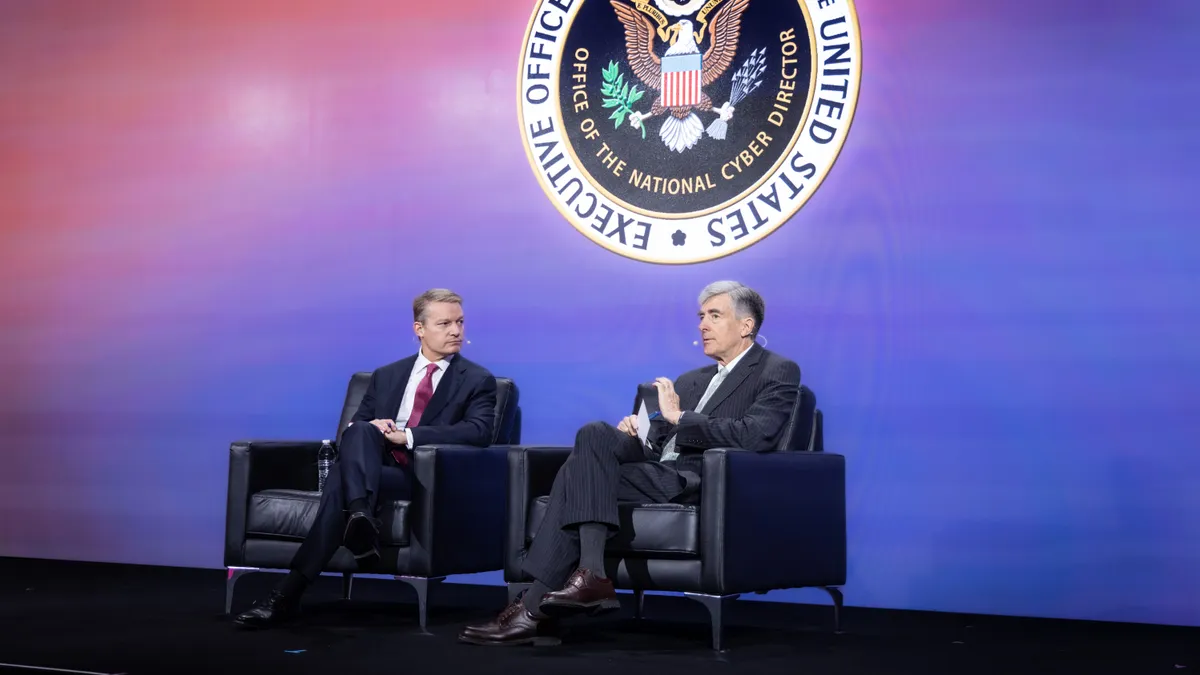 Two men sit on stage in front of a logo of the executive office of the president national cyber director.