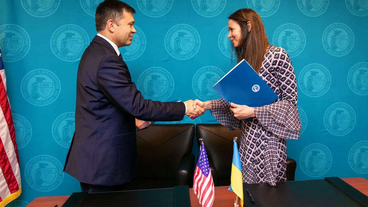 A man and a woman shake hands in front of a desk that has flags from the U.S. and Ukraine. The people are in front of a blue background with CISA logos.